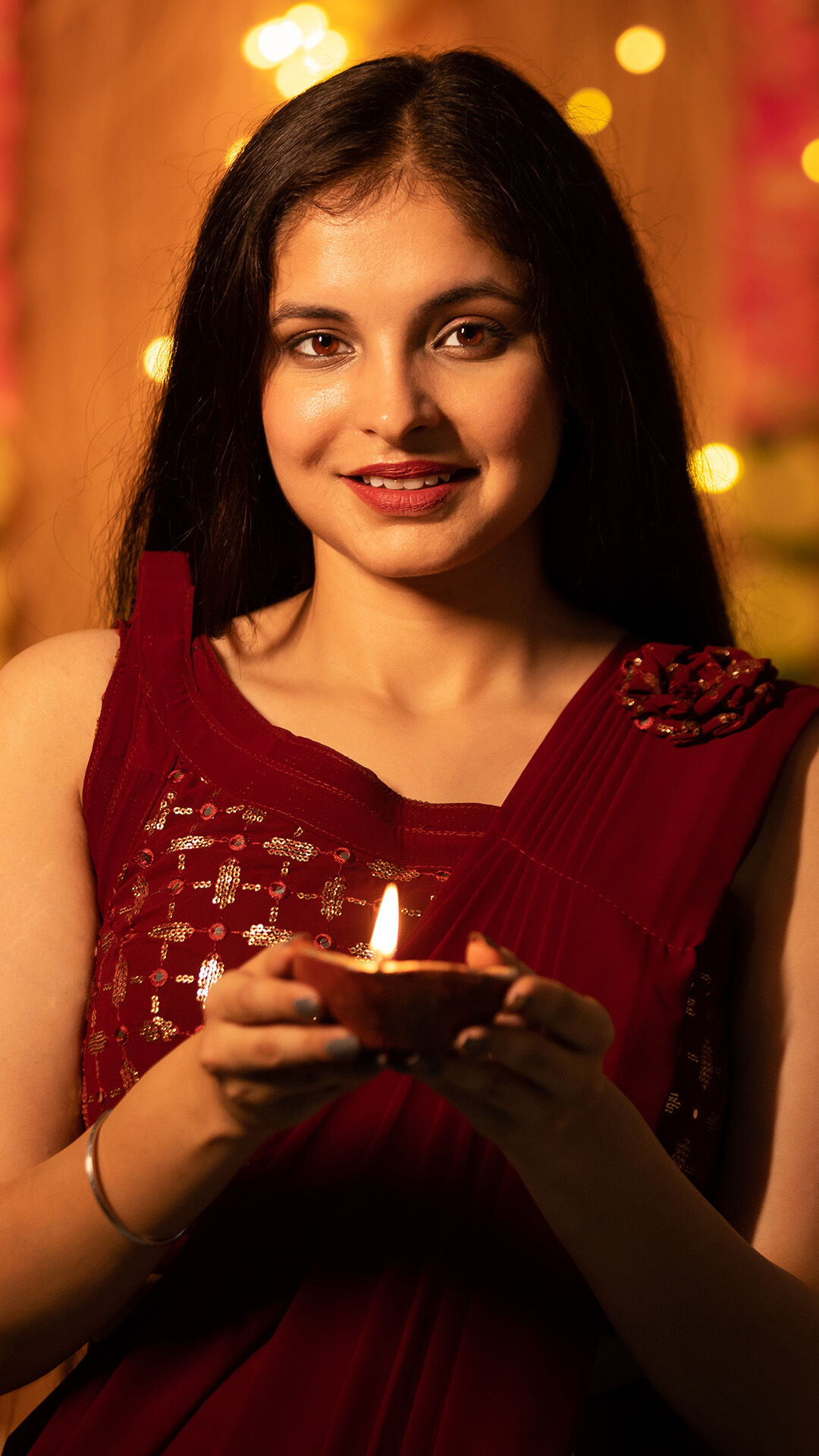 A woman in a red festive outfit holding a glowing diya, a symbol of light during Diwali.